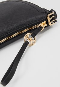 Clutch de cuero negro con acabado texturizado, cierre de cremallera dorado y colgante con el logo de MK en una correa negra. Forma redondeada y diseño elegante.