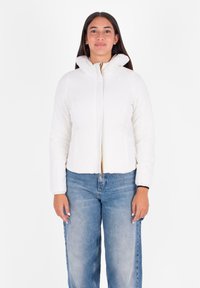 Vrouw met lang donker haar, gekleed in een witte gewatteerde jas en een spijkerbroek, die rechtop staat tegen een plain witte achtergrond.
