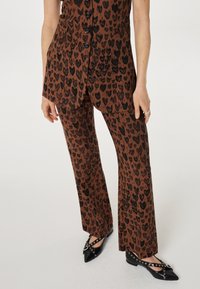 Twee-delig outfit bestaande uit een bruine stof met zwarte hartvormige leopardvlekken; inclusief een knopenvest en uitlopende broek, gecombineerd met zwarte schoenen.