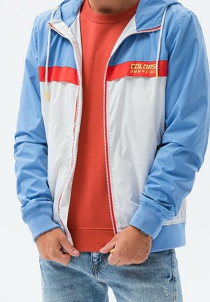 Homme portant une veste zippée bleue, blanche et rouge sur un t-shirt orange et un jean bleu clair, ajustant la fermeture éclair de la veste.