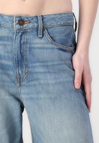 Lyseblå denimjeans med høyt liv, knappeåpning og sidelommer. Har synlig søm og en lett falmet tekstur.