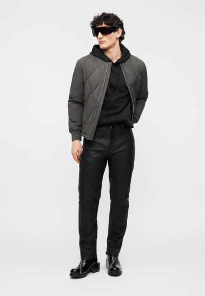 Άνδρας με μαύρη φούτερ με κουκούλα, γκρι καπιτονέ μπουφάν, μαύρο δερμάτινο παντελόνι, μαύρα μπότες με τακούνι και υπερμεγέθη γυαλιά ηλίου, στέκεται μπροστά σε απλό φόντο.