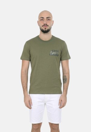 Uomo con barba e tatuaggio che indossa una t-shirt verde oliva "Classic Arts" e pantaloncini bianchi, in piedi davanti a uno sfondo chiaro e uniforme.