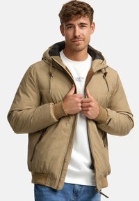 Unge mand iført beige hættetrøje over hvid sweatshirt og blå jeans, holder jakkezipperen med begge hænder.