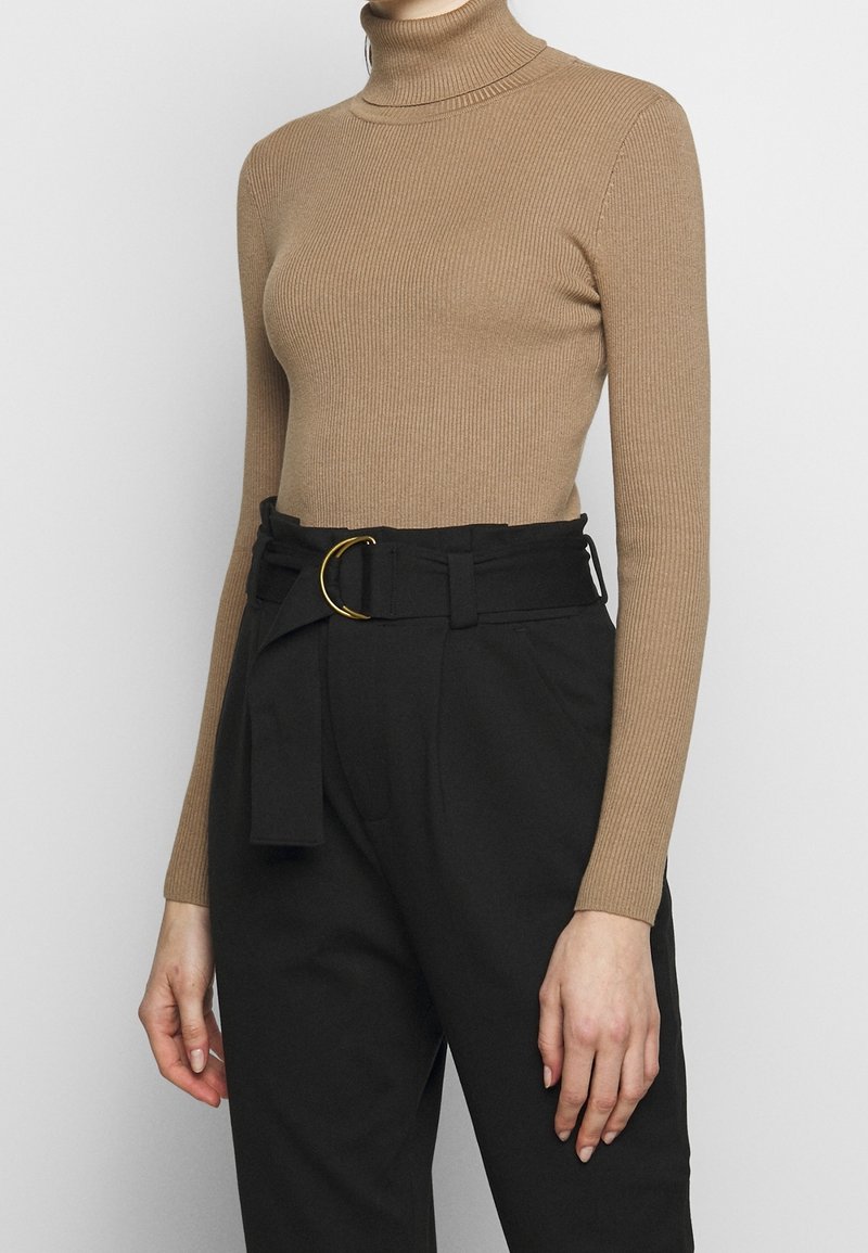 Personne portant un pull col roulé côtelé beige ajusté, rentré dans un pantalon noir taille haute avec une ceinture large et une boucle dorée.