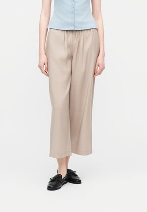 Γυναίκα που φοράει μπεζ παντελόνι με φαρδιά μπατζάκια έως τον αστράγαλο, ανοιχτό μπλε αμάνικο πουκάμισο με κουμπιά και μαύρα δερμάτινα μοκασίνια, στέκεται μπροστά σε απλό φόντο.