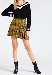 Schwarzer Pullover mit weißem V-Ausschnitt und Branding an den Ärmeln, kombiniert mit einem gelben karierter, plissierter Rock mit Schnallen. Schwarze Knöchelstiefel runden den Look ab.