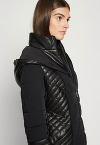 Crna zimska jakna s puhovima sa kvadratnim uzorkom s jedne strane, kontrastni glatki materijal koji podsjeća na kožu na kapuljači i gornjem dijelu tijela. Čičak zatvarač.