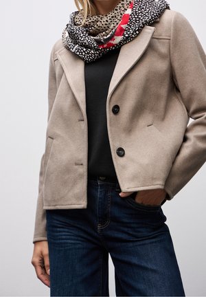 Femme portant une veste beige boutonnée sur un haut noir, un jean bleu foncé et une écharpe multicolore à motifs autour du cou, la main dans la poche.