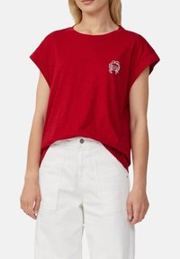 Vrouw in een rood T-shirt met korte mouwen en een klein wit krabontwerp op de borst, gestopt in hoge witte broeken.