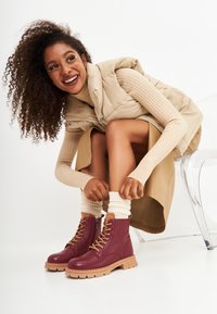 Botas de tobillo de cuero burdeos con cordones beige y una suela gruesa de color marrón claro. Llevadas con calcetines blancos de canalé y un atuendo neutro.