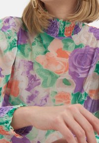Blouse légère en tissu transparent à motif floral avec un col haut froncé et des poignets dans des tons de violet, vert et corail. Tissu texturé au motif éclatant.