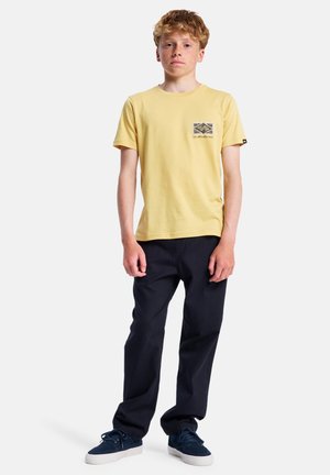 Geel katoenen T-shirt met een contrasterende gemusterde zak, gecombineerd met marineblauwe broek en donkere sneakers. Model staat rechtop tegen een effen achtergrond.
