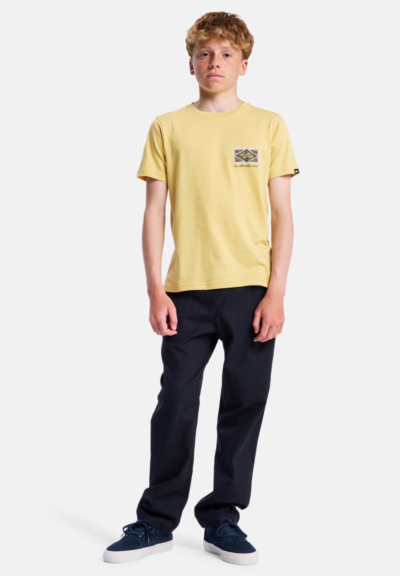 Žluté bavlněné tričko s kontrastní patterned kapsou, spárované s námořnickými kalhotami a tmavými teniskami. Model stojí vzpřímeně na jednobarevném pozadí.