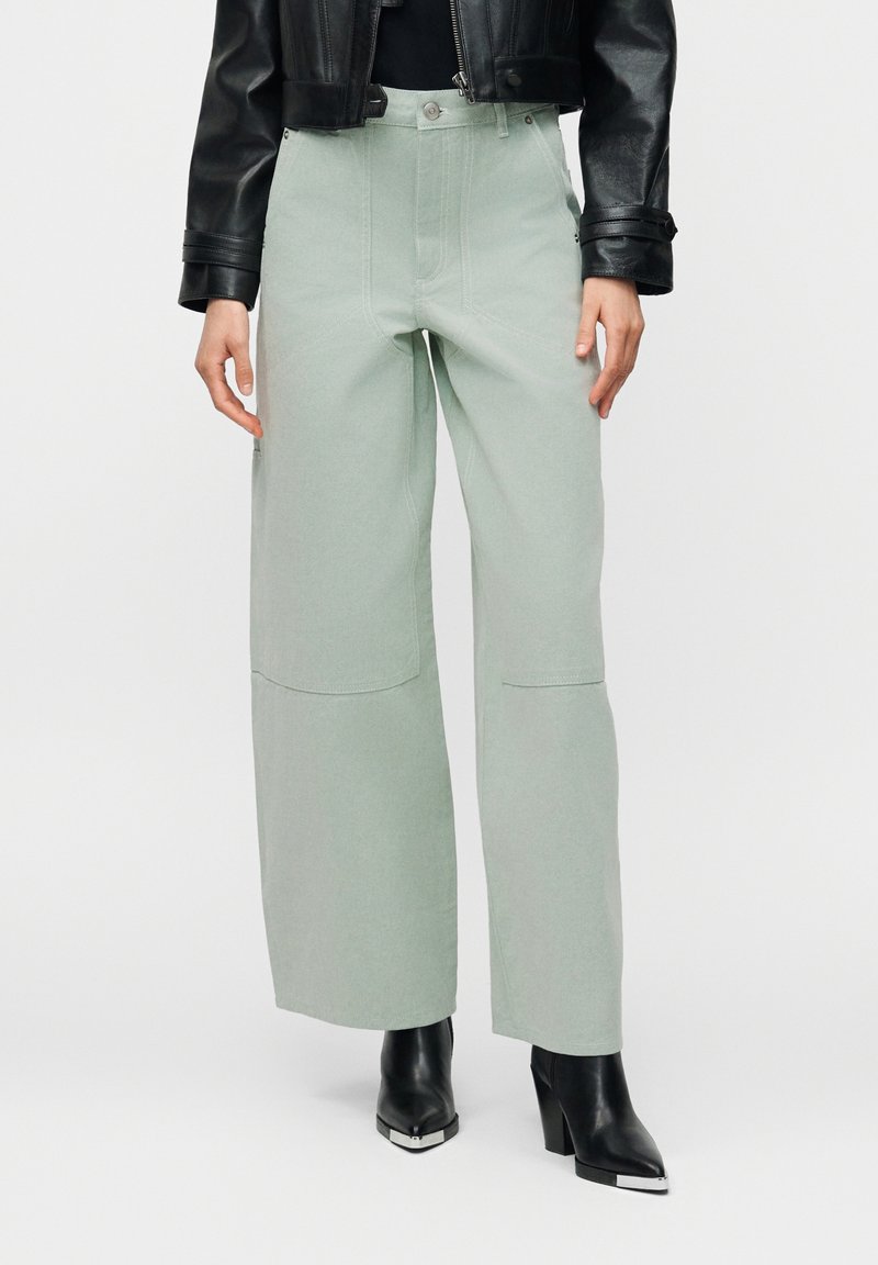 Γυναίκα που φοράει ανοιχτό πράσινο παντελόνι με φαρδύ πόδι και τσέπες, μαύρο κοντό δερμάτινο μπουφάν και μαύρα κοντά μποτάκια με τακούνι και ασημένιες μύτες.