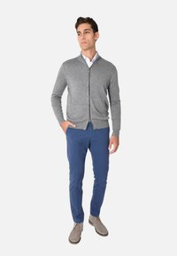 Cardigan grigio con zip sopra una camicia bianca, abbinato a pantaloni blu e scarpe beige. Il cardigan ha una texture liscia e un design aderente.