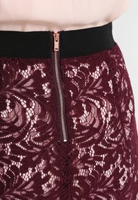 Burgundy spetskjol med ett blommönster, svart elastisk midja och en roséguldfärgad dragkedja baktill. Texturen verkar ömtålig.