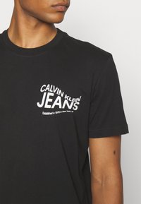 Black cotton T-shirt with short sleeves. Features white text: "CALVIN KLEIN JEANS" and "Established in 1978 in New York, NY" in varying fonts.