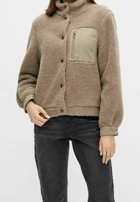 Donna che indossa una giacca in sherpa beige con bottoni a pressione e una tasca sul petto con zip, abbinata a jeans neri.
