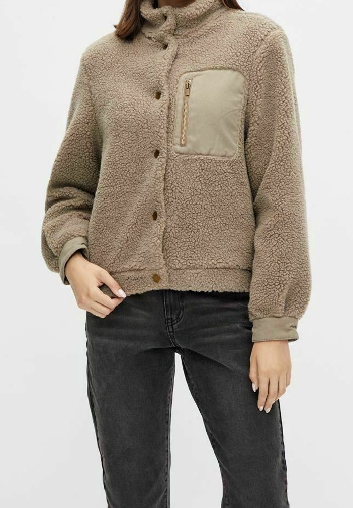 Donna che indossa una giacca in sherpa beige con bottoni a pressione e una tasca sul petto con zip, abbinata a jeans neri.