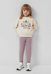 Kremowy sweatshirt z kreskówkowym wzorem graficznym; tekst "PRZYJAŹŃ to najczystsza miłość." W zestawie z lawendowymi legginsami i białymi sneakersami.