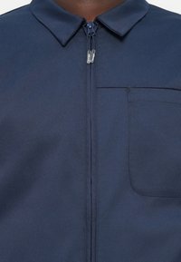 Chaqueta de cremallera azul marino con un pequeño collar, bolsillo frontal y textura suave y mate; la tela parece duradera y ligera.