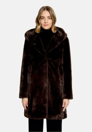 Frau mit schulterlangem blonden Haar, die einen dunkelbraunen Kunstpelzmantel mit Kapuze über einem schwarzen Rollkragenpullover und Hose trägt.