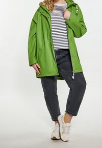 Jasně zelená nepromokavá bunda s kapucí, stříbrnými zipy a patentkami. Nositá přes černobílý pruhovaný svetr a tmavé džíny.