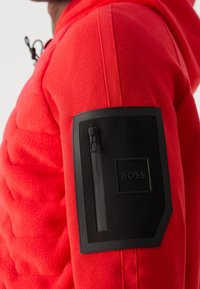 Närbild av en röd jackärm med en svart rektangulär patch som har en vertikal dragkedja och upphöjd text "BOSS".