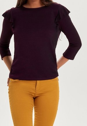 Donker paarse aangesloten top met ruffles aan de mouwen, ronde halslijn en driekwart mouwen, gecombineerd met mosterdgele broek.