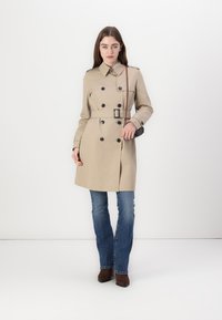 Beige trenchcoat met dubbelgeknoopte ontwerp, zwarte knopen, riem op de taille en schouderepauletten, gecombineerd met blauwe jeans en bruine laarzen.
