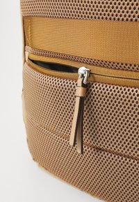 Brauner Netzstoff-Rucksack mit strukturierter Oberfläche, ausgestattet mit einer vorderen Reißverschlusstasche und einem Lederreißverschluss-Zugbandakzent.