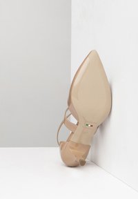 Nude patent leather high-heeled shoe with a pointed toe, ankle straps, and a tapered heel. Features branding and size details on the sole.