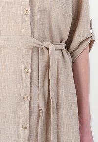 Robe chemise en lin beige avec une taille à nouer, manches courtes retroussées, détail de boutons sur le devant et poche poitrine. Tissu doux et texturé.