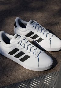 Zapatillas Adidas blancas con rayas negras y cordones, colocadas sobre una superficie gris texturizada, mostradas desde un ángulo lateral y superior.