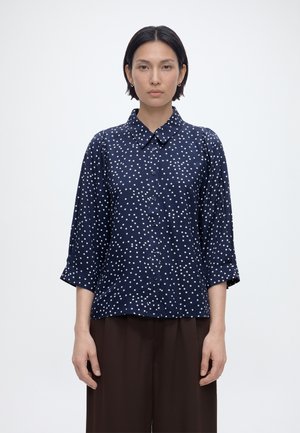 Mujer de cabello corto y oscuro lleva una camisa de manga larga azul marino con lunares blancos y pantalones anchos oscuros, de pie contra un fondo liso.