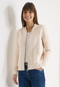 Femme souriante, portant une veste beige zippée sur un haut blanc avec un jean bleu, debout contre un mur blanc, une main dans la poche.