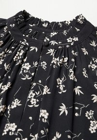 Tissu noir avec petit imprimé floral blanc, avec un col à volants froncés et une fermeture par bouton avec boucle en tissu au niveau de l'encolure.