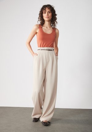 Rode tanktop met smalle banden, gecombineerd met brede beige broek, voorzien van een riem en voorplooien, aangevuld met zwarte platte schoenen.