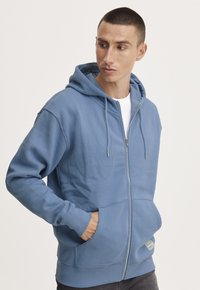 Sudadera con cremallera de color azul claro, hecha de una tela suave, con capucha ajustable, bolsillos frontales y puños y dobladillo de canalé.
