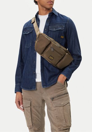 Uomo che indossa una camicia di jeans blu, pantaloni cargo beige e una borsa a tracolla verde oliva con molteplici tasche con cerniera.