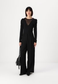 Svart långärmad topp med genomskinlig V-ringning, tillsammans med vida svarta byxor. Modellen håller en svart handväska. Enkel monokrom design.