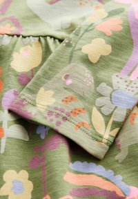 Zielony materiał z kolorowymi wzorami kwiatowymi i abstrakcyjnymi, z widocznym szwem i marszczeniami.