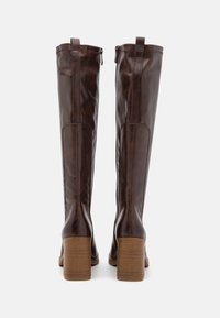 Ruskean nahkainen polvipituinen saappaat, joissa on sileä pinta, vetoketju takana ja paksu korko. Korko on puumaisen viimeistelyn omaava.