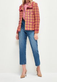 Veste à carreaux roses et orange avec un col pointu, deux poches avant, associée à un jean bleu droit et des talons hauts couleur nude.