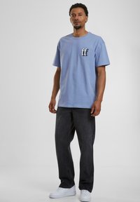 Lys blå bomull T-skjorte med korte ermer, med en svart logo på venstre bryst. Kombinert med mørke grå løse jeans og hvite joggesko.
