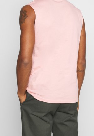 Homme portant une chemise rose pâle sans manches et un pantalon gris foncé, mains dans les poches, montrant le dos et des tatouages sur les bras.