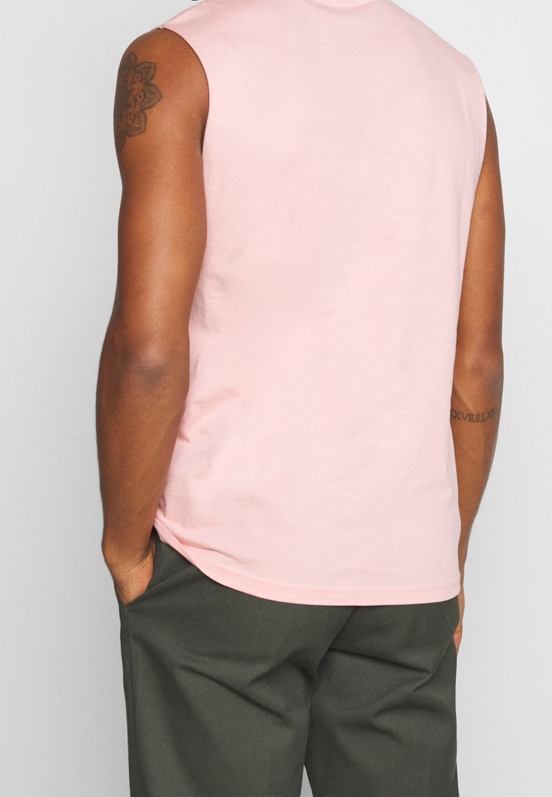 Homme portant une chemise rose pâle sans manches et un pantalon gris foncé, mains dans les poches, montrant le dos et des tatouages sur les bras.