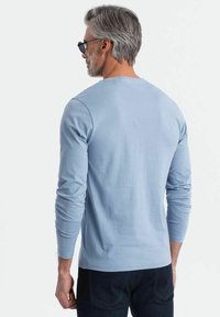 Camicia azzurra a maniche lunghe realizzata in tessuto morbido, con scollatura rotonda e polsini aderenti, abbinata a jeans scuri. Vista da dietro.