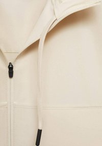 Beige zip-hoodie med en glat stoftekstur, der har en sort lynlås og snøre detaljer med sorte spidser.